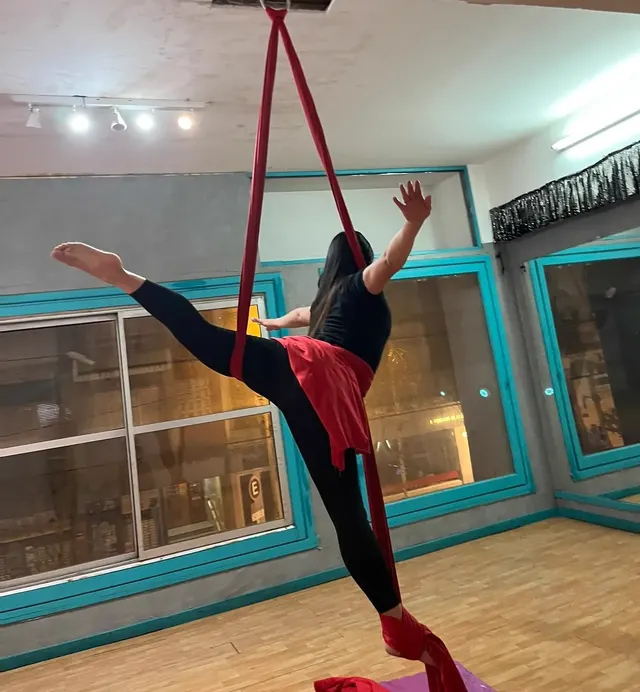 Alumna en el Estudio Melani Mitchell suspendida en la tela roja, con una pierna estirada hacia atrás y los brazos abiertos frente a las ventanas turquesa del estudio
