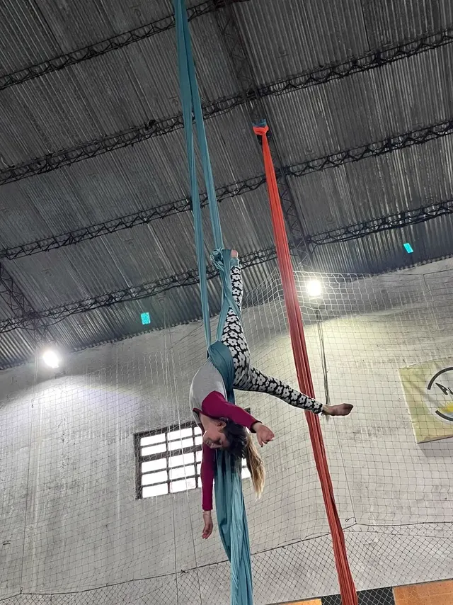 Alumna invertida en la tela turquesa del Gimnasio Alma, con las piernas abiertas y el pelo hacia el piso, bajo el techo de chapa