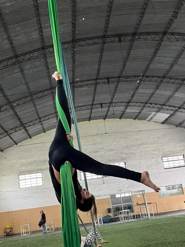 Alumna invertida en la tela verde del Gimnasio Alma, con una pierna enroscada en la tela y la otra estirada, en la cancha de césped sintético con el cartel del gimnasio al fondo