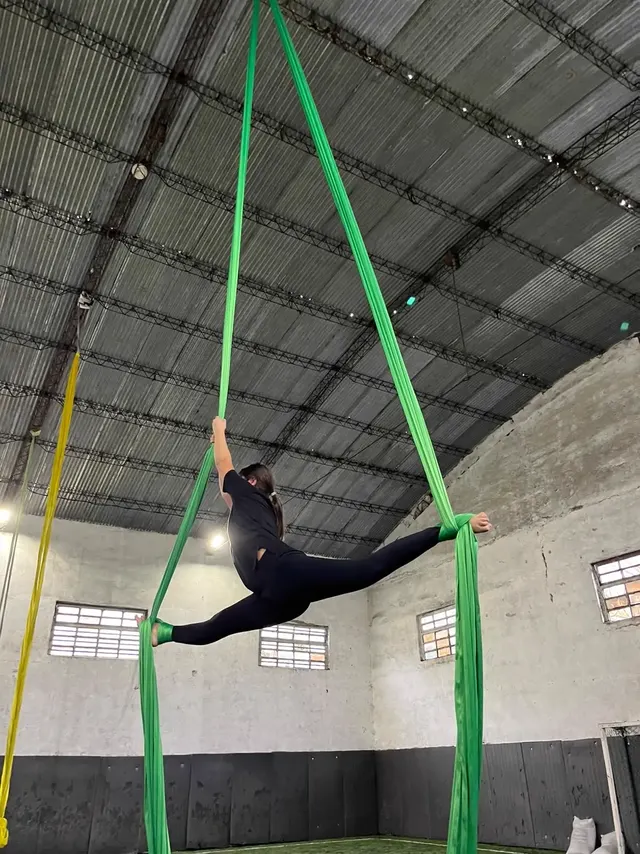 Alumna en el Gimnasio Alma en split en el aire entre las dos caídas de la tela verde, bajo el techo parabólico de la cancha techada