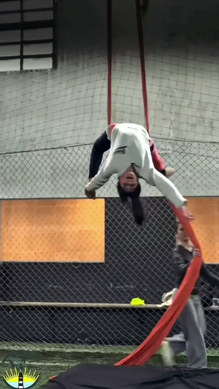 Clase de acrotela en el Gimnasio Alma: una alumna baja en figura sobre la tela roja en la cancha techada, mientras el resto del grupo entrena en las otras telas, los lunes y viernes a las 16:15